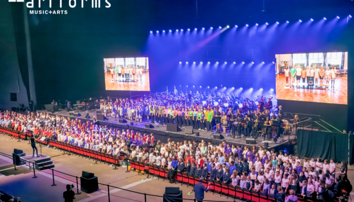 A wide shot of an orchestra and large school choir on and in front of the Leeds Arena stage. Lots of spotlights and two screens either side of the stage showing a smaller school choir.
