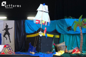 Set for the YAMSEN flies to Neverland event. There is a blue cloth backdrop with a painted boat in the centre, a silhouette of Peter Pan on the left and a palm tree on the right.
