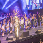 Lots of primary school children on the Leeds arena stage with one arm in the air following the conductor at the front. Blue spotlights behind the choir and fireworks at the front of the stage.