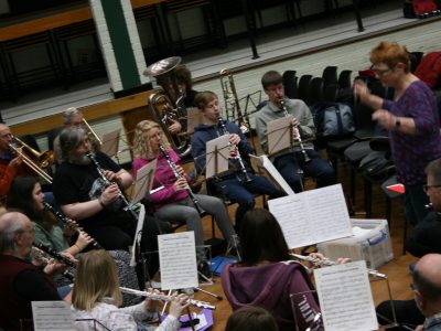 Concert band playing at a music centre in a school hall. Conductor is standing at the front with her hands moving.