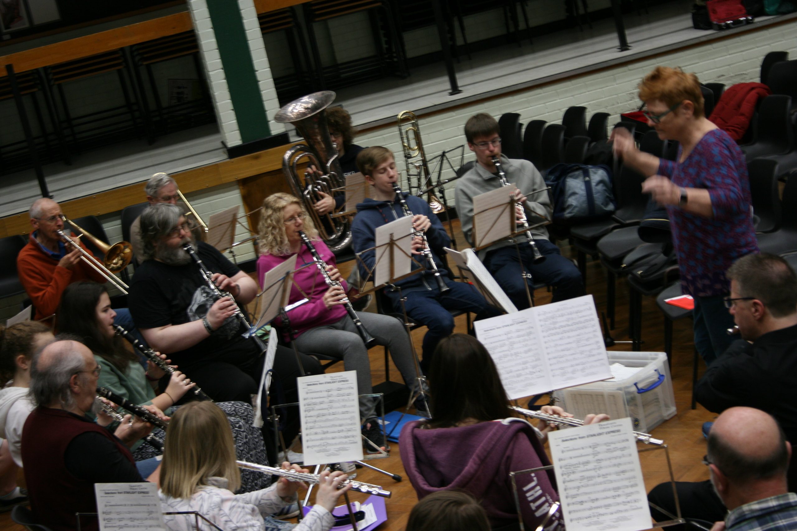 Concert band playing at a music centre in a school hall. Conductor is standing at the front with her hands moving.